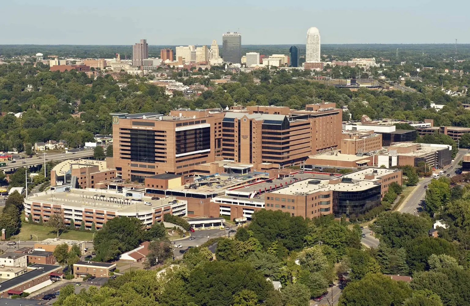 Atrium Health Wake Forest Baptist Medical Center in Winston-Salem, featuring modern hospital buildings surrounded by greenery with the city skyline in the background.